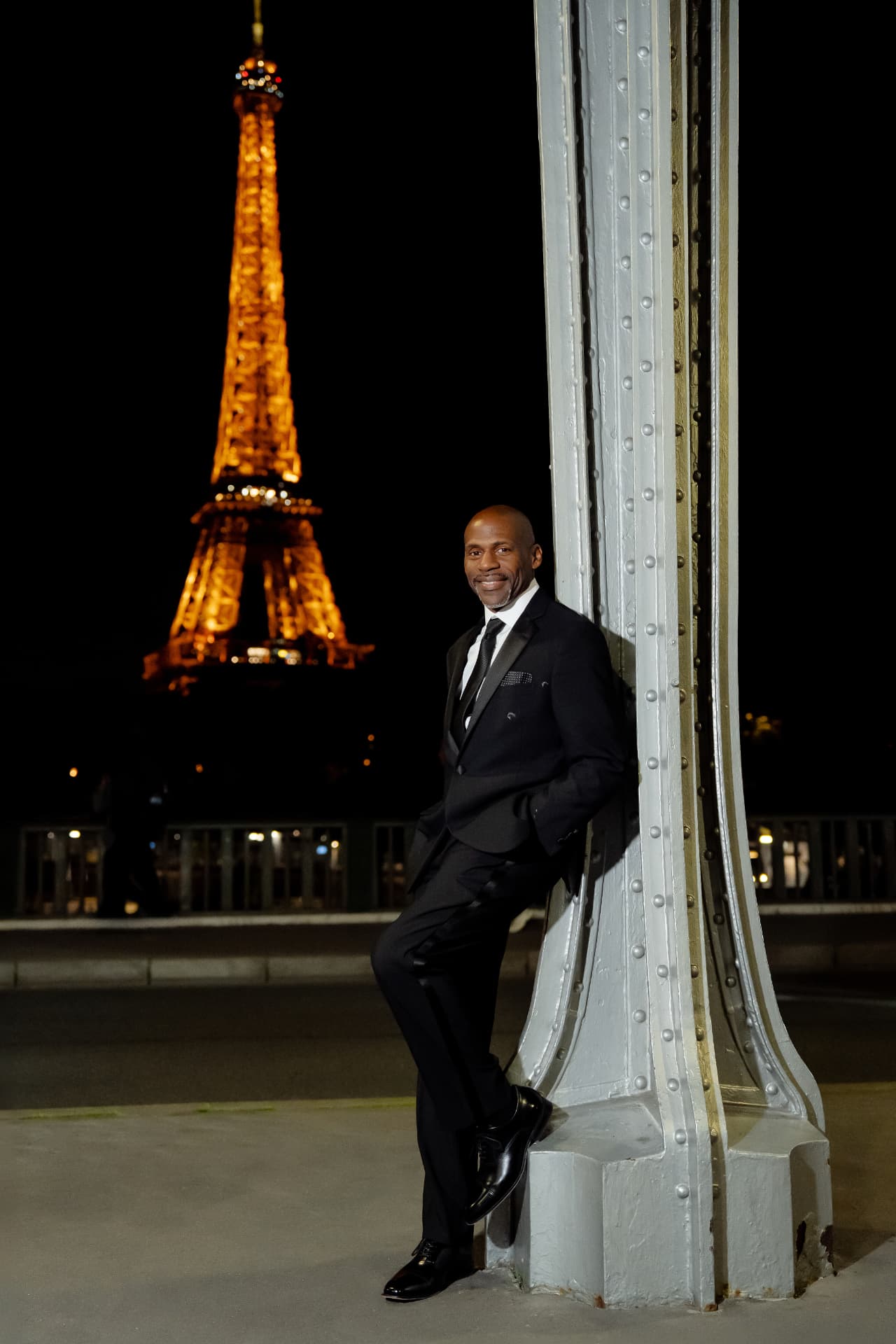 Eone in a black tuxedo leaning against a bridge column with the Eiffel Tower behind him
